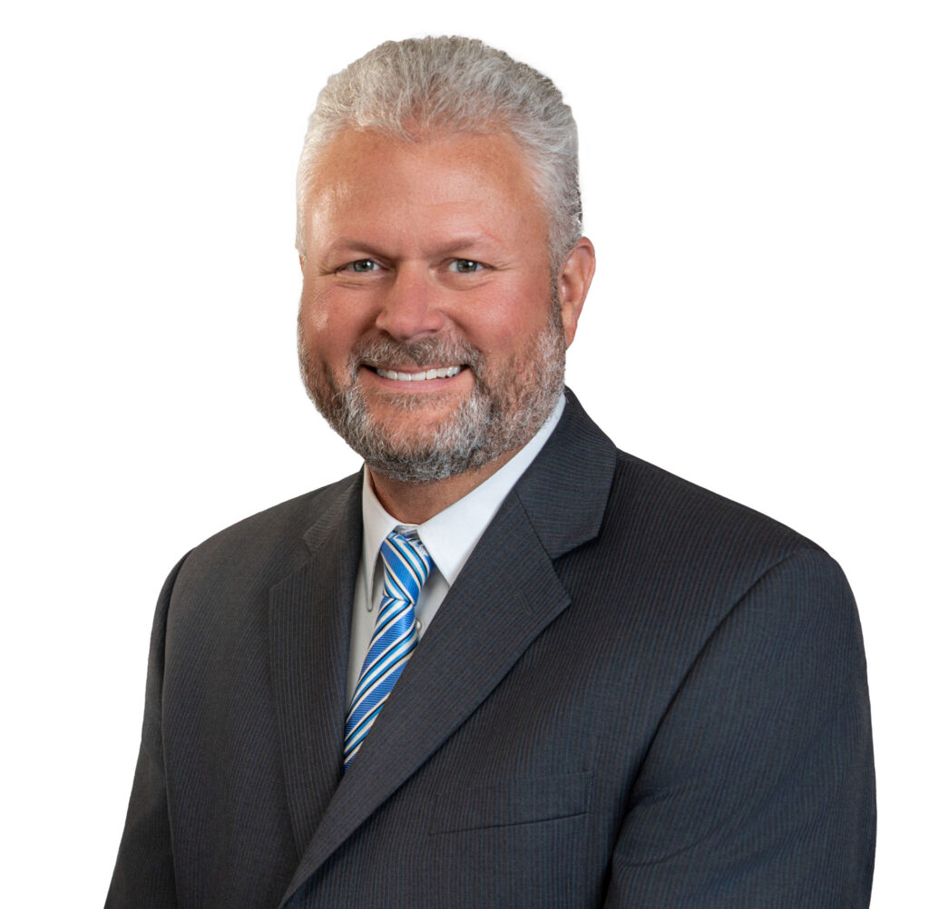 A smiling middle-aged man with gray hair and a beard, wearing a gray suit, white shirt, and blue-striped tie.