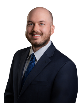 A man with a beard and shaved head wears a navy suit, light blue shirt, and patterned blue tie. He is smiling, facing slightly to the left, with a plain white background.