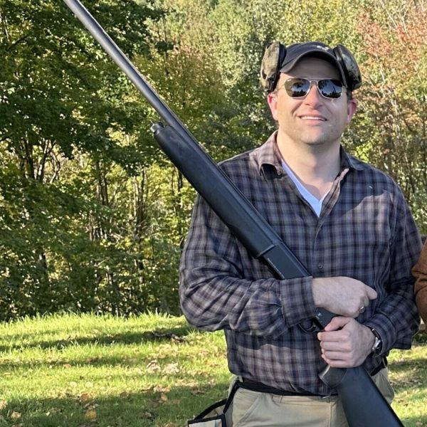 A man wearing sunglasses, a plaid shirt, khaki pants, and a camouflage cap stands outdoors on grass, holding a large black shotgun vertically—his stance confident as if ready for anything, much like seasoned criminal defense attorneys. Trees with green and autumn leaves are in the background.
